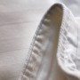 Close-up image of the corner of a white bed sheet or fabric, showing its hem stitching and woven texture. Made from luxurious Mulberry Silk, the image highlights the intricate details of the stitching and the silky fabric material.