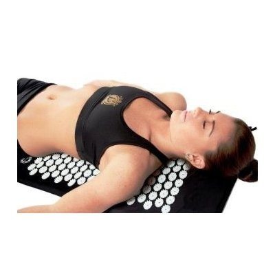 A person wearing a black sports bra and black shorts lies on an acupressure mat with white circular pressure points. The person has their eyes closed and appears to be relaxing or meditating.