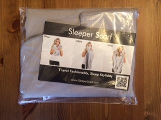 A packaged Sleeper Scarf on a wooden surface. The packaging shows three images demonstrating how to wear, inflate, and sleep with the Sleeper Scarf. The text reads "Travel Fashionably, Sleep Stylishly" and provides a website URL along with a QR code.