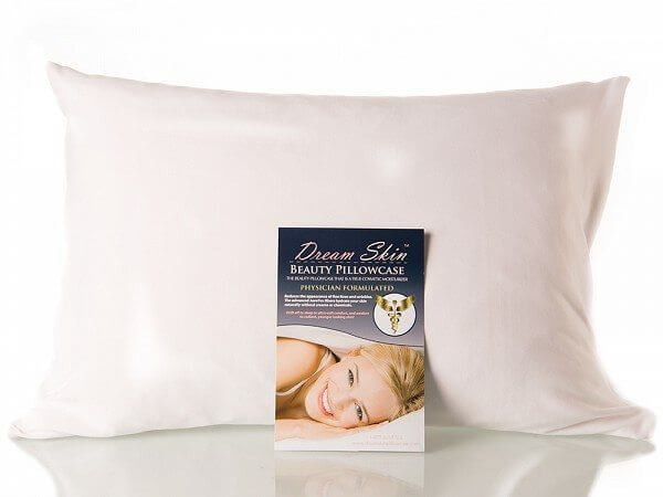 A large white pillow stands upright against a light background. In front of it is a product package for the "Dream Skin Pillowcase." The package features an image of a smiling woman lying down and text detailing the beauty benefits of the pillowcase.