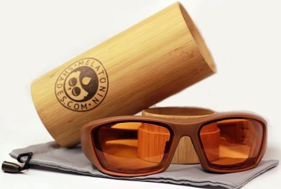 Wooden-framed amber-tinted Melatonin Shades are displayed in front of a cylindrical bamboo case adorned with a logo of a sleeping face and the words "THE ME LATONIN D ES CO M.N." on it. The sunglasses and case rest on a gray drawstring pouch.