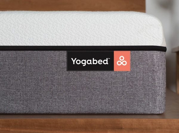A close-up view of a mattress from the innovative Mattress Startup "Yogabed," featuring a black and red label on the lower gray fabric section. The upper section of the mattress is white, with a wooden surface backdrop completing the scene.