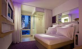 Modern hotel room at Yotel with a cozy double bed, white linens, and plush pillows. The room features a wall-mounted TV, ambient purple lighting, a bathroom with a glass shower, and a large window showcasing a nighttime city view with illuminated buildings.