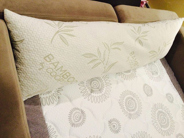 A large bamboo pillow rests on the arm of a sofa. The Shredded Memory Foam Pillow cover is white with a bamboo leaf print and the word "BAMBOO" printed on it. Below the pillow, a white quilt with a circular pattern design is draped over the sofa cushions.