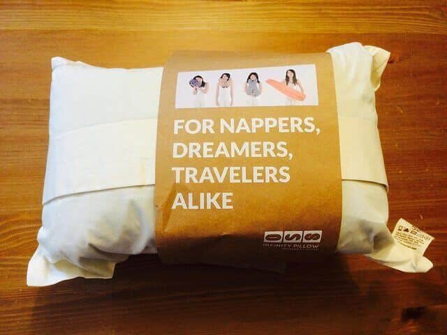 A white, plush pillow is shown wrapped in brown paper packaging. The packaging features the text "FOR NAPPERS, DREAMERS, TRAVELERS ALIKE" and displays four small images of individuals using the Huzi Pillow in different ways. The brand name "ONE INFINITY PILLOW" is also visible.