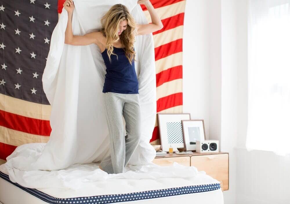 A woman in pajamas stands on a WinkBeds mattress while holding a white sheet above her head. Behind her, a large American flag is hung on the wall. The room is filled with bright natural light, and in the background, there are framed pictures and a small speaker on a dresser.