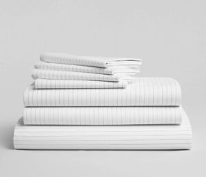 A neatly folded stack of white bed linens with thin grey vertical stripes is arranged against a plain grey background. The stack includes pillowcases, sheets, and a larger folded item, possibly a duvet cover. The fabric appears clean and crisp—a perfect setup for an excellent Brooklinen review.