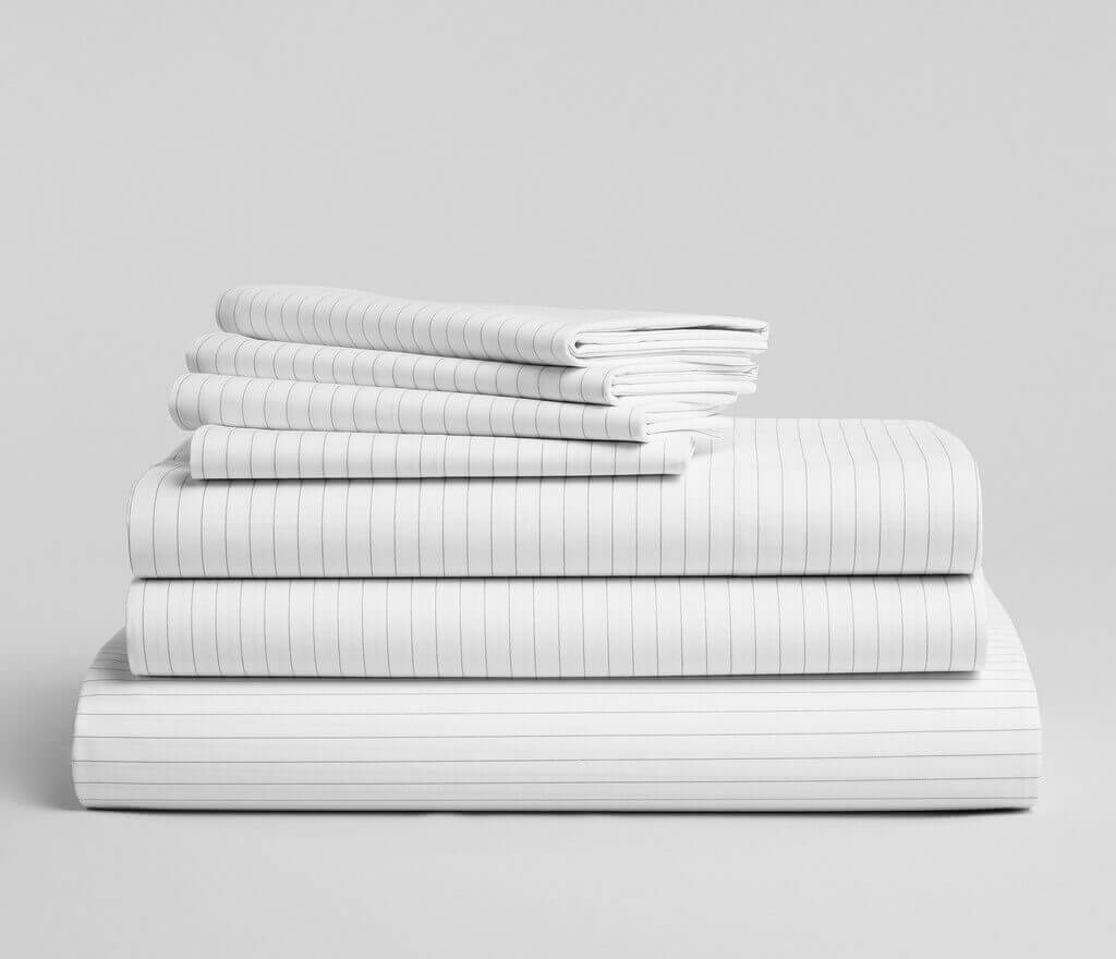 A neatly folded stack of white bed linens with thin grey vertical stripes is arranged against a plain grey background. The stack includes pillowcases, sheets, and a larger folded item, possibly a duvet cover. The fabric appears clean and crisp—a perfect setup for an excellent Brooklinen review.