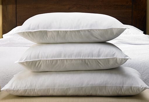 Three neatly stacked white pillows are placed on a bed with a wooden headboard. In the background, there are white bed linens, contributing to a clean and comfortable bedroom setting. This could easily be a scene from a Hampton Inn Pillow Review, showcasing their attention to comfort and style.