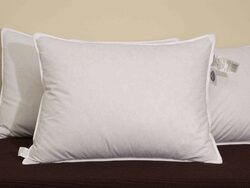 A white Marriott pillow rests on a dark surface with another pillow partially visible in the background. Both pillows feature white piping along the edges and a tag attached to the visible one. The backdrop is a neutral beige color, highlighting the elegance of the Marriott bedding.