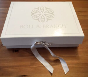 A white Boll & Branch gift box with a decorative logo on the lid is placed on a wooden surface. The box is neatly tied with a light gray ribbon, forming a bow on the front.