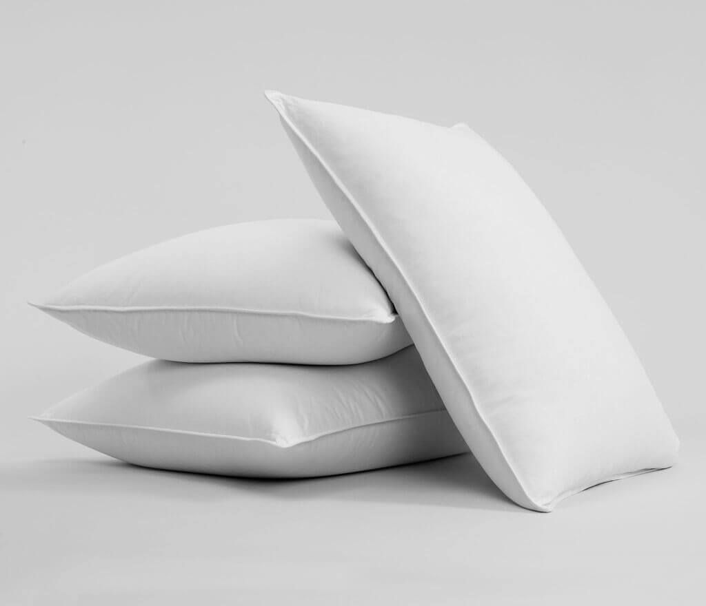 Three Brooklinen Pillows are stacked against a plain gray background. Two pillows are laid flat on the bottom, and the third pillow is balanced upright on top of them.