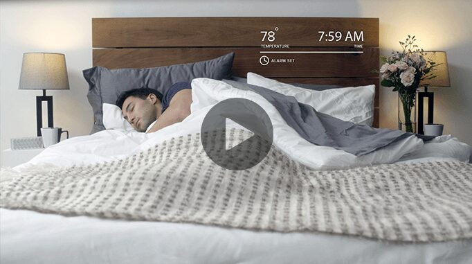 A man sleeps peacefully in a modern bedroom on a white bed with gray and white blankets. On the headboard, a digital display shows the temperature as 78°F, the time as 7:59 AM, and that an alarm is set. Two lamps and a flower vase are on bedside tables nearby.