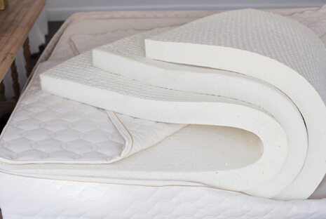 A tri-fold foam mattress topper partially unfolded and placed on a white mattress. The topper has a textured surface and appears to be made of a thick, cushioned material for added comfort.