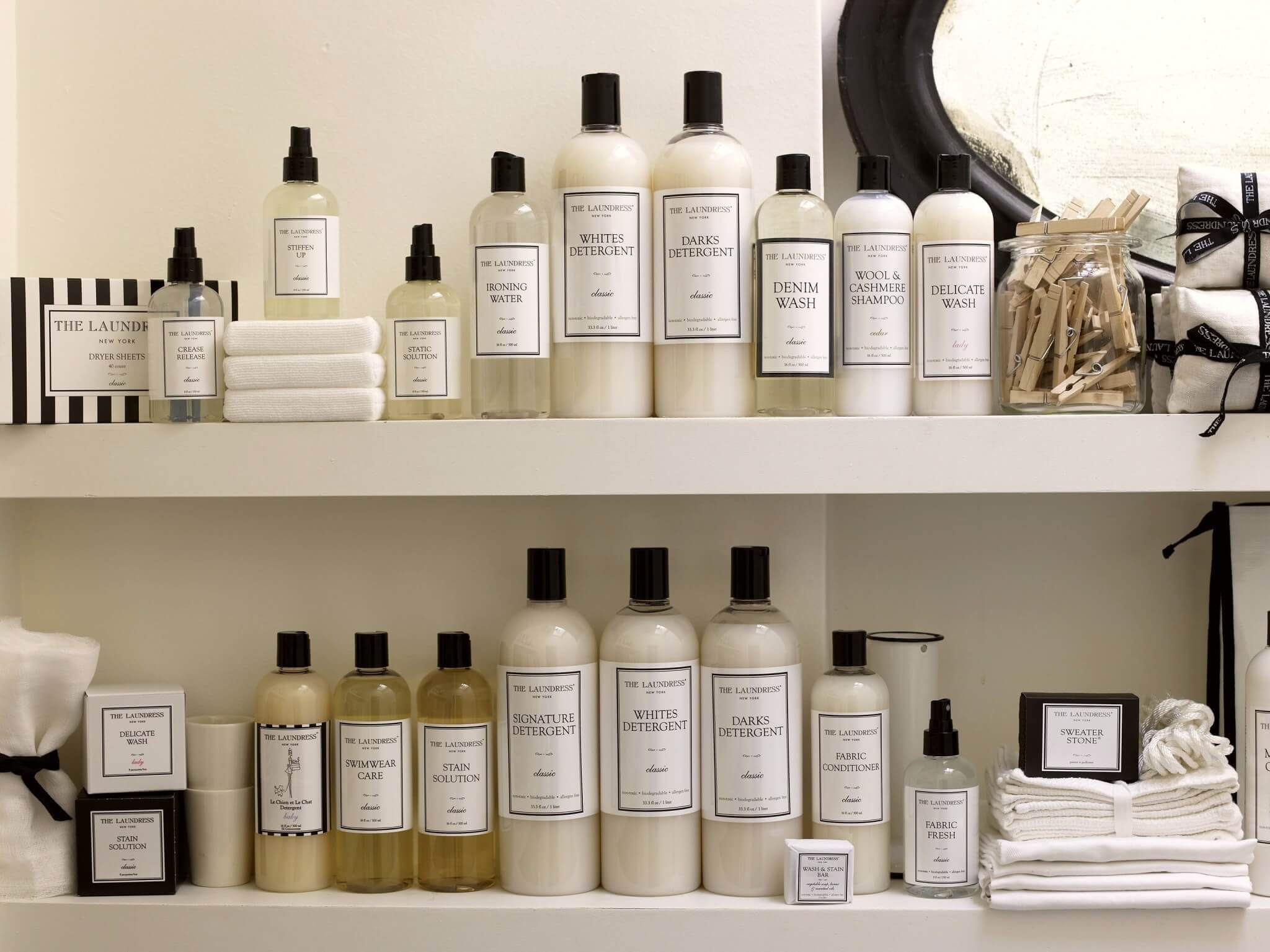 A shelf filled with various white and clear bottles and containers of cleaning and laundry products from The Laundress. Items include detergents, fabric sprays, and accessories such as small stacked towels and clothespins in a glass jar, neatly organized—truly deserving of a glowing Laundress review.