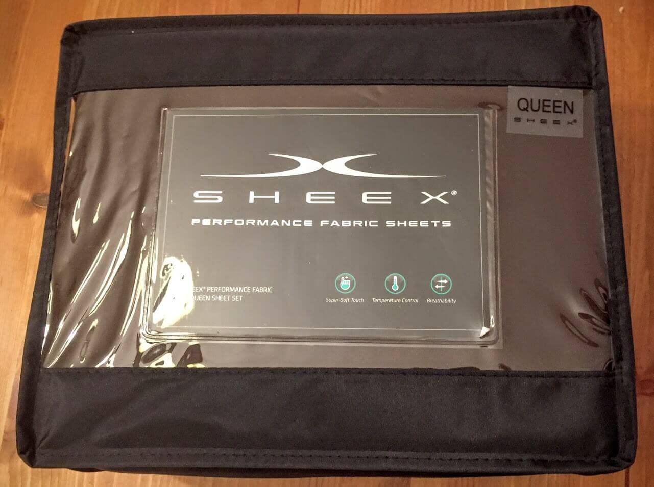 A packaged set of Sheex Performance Fabric Sheets in queen size lays on a wooden surface. The dark packaging has a clear window showing the product label, which highlights features like temperature control, super-soft touch, and breathability. For more details, check out a comprehensive Sheex Review online.