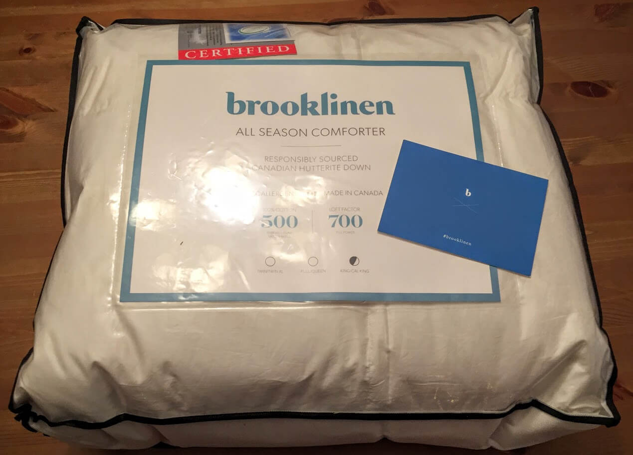 A packaged Brooklinen All Season Down Comforter is placed on a wooden surface. The label indicates it is responsibly sourced Canadian Hutterite down with a 500 fill power and 700 thread count. A blue card with the Brooklinen logo is next to the package.