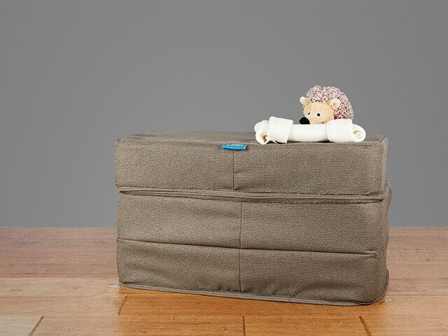 A rectangular, green storage pouch sits next to a foam dog bed on a wooden table against a gray background. On top of the pouch, there's a plush toy hedgehog holding a dog bone toy in its paws.