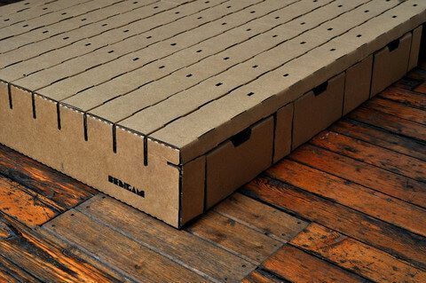 A Bedigami cardboard platform bed with storage drawers integrated into the base sits on a rustic wooden floor. The bed and drawers are made from corrugated cardboard, featuring cutout handles for easy access to the storage compartments.