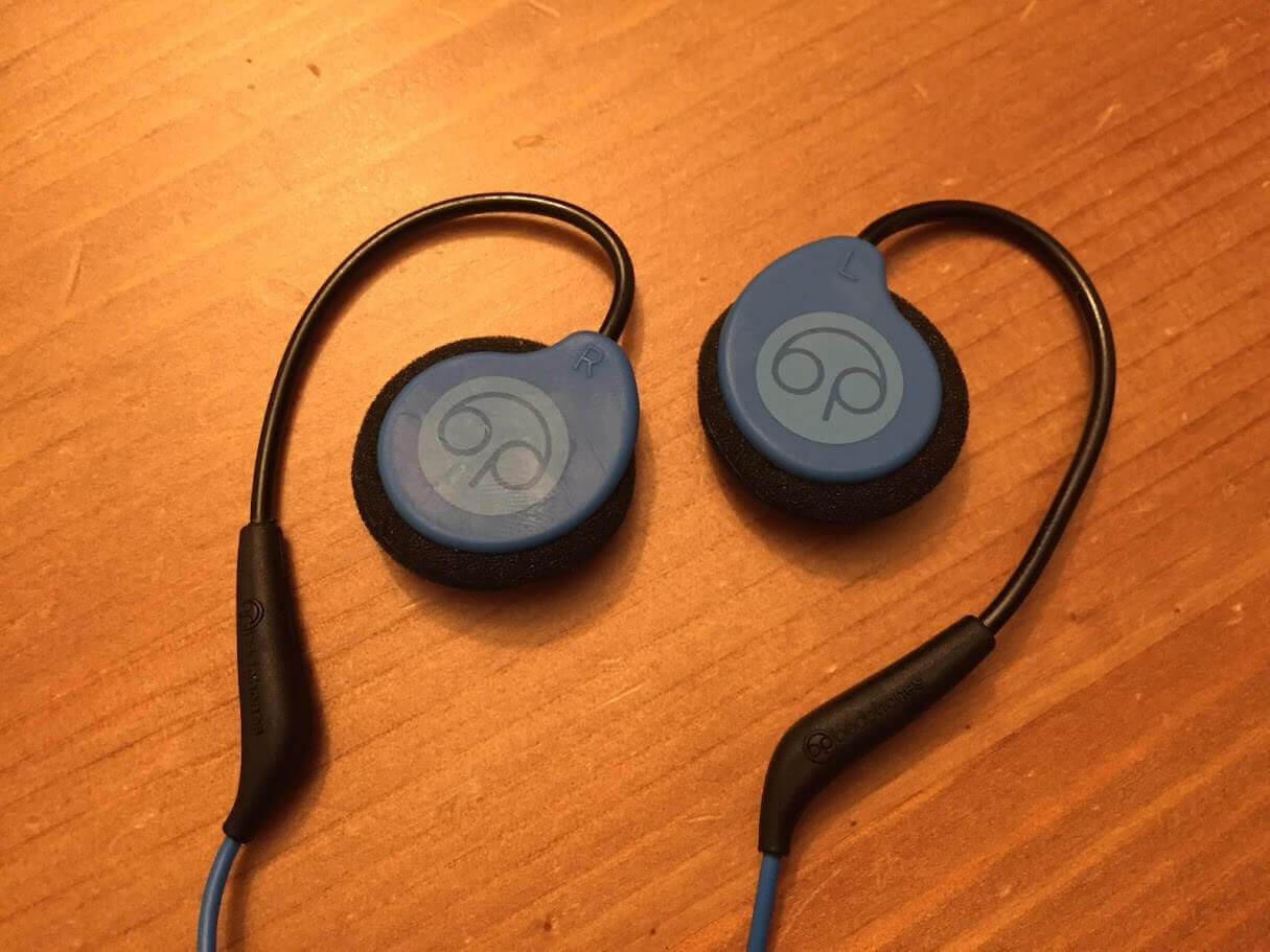 A pair of blue wireless on-ear sports headphones, akin to bedphones in comfort, with black ear hooks sits on a wooden surface. The headphones feature a blue circular design with the company logo in the center and black padding around the edges for added comfort.