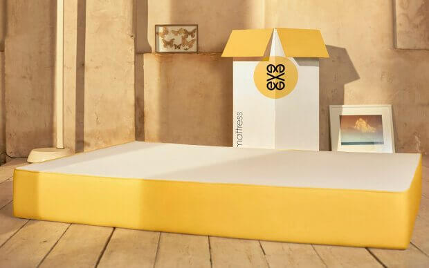 A yellow-and-white Eve Mattress is placed on a wooden floor in a sunlit room. Behind the mattress, a large open box is visible, with the brand logo and the word "mattress" on it. A framed picture and a butterfly decor are positioned on the floor and wall, respectively.