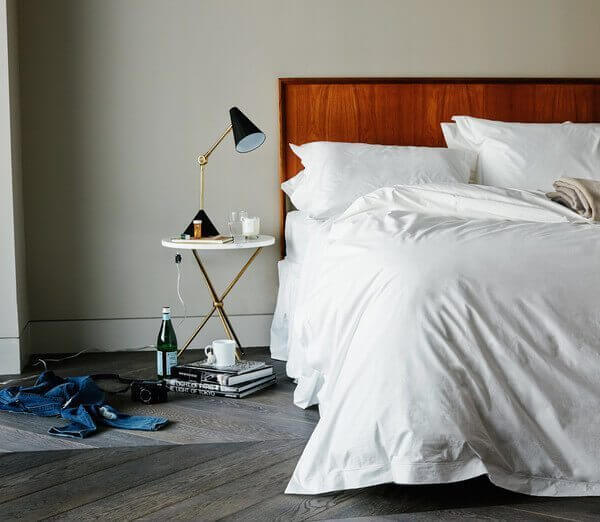 A neatly made bed with Snowe white linens sits against a light gray wall. A wooden headboard is visible. To the left, a small round side table holds a modern black lamp, a glass of water, and a candle. On the floor, a pair of jeans, a water bottle, and books are scattered.