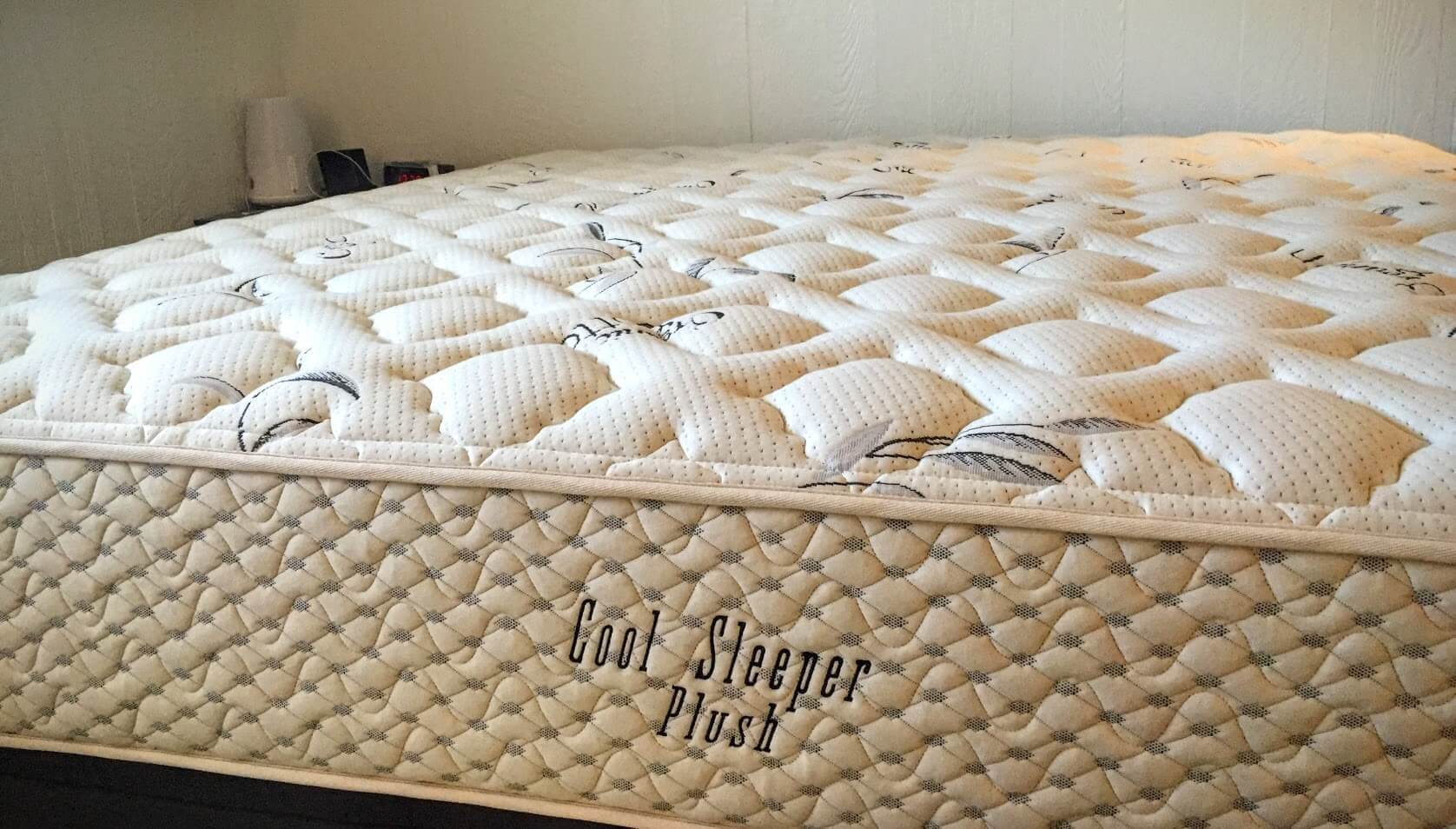 A plush Honest Beds mattress with textured quilting and lightly stitched floral designs, labeled "Cool Sleeper Plush" on the side, is shown in a bedroom setting. A small bedside lamp is visible in the background.