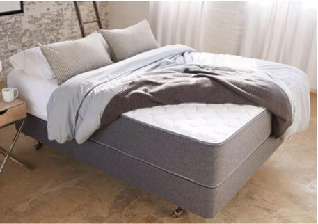 A neatly made bed with gray pillows and a matching gray comforter on an Aviya Mattress. The bed frame is gray, and the bed is positioned in a minimalistic room with a rustic wall, a small side table, and a window with sheer white curtains.