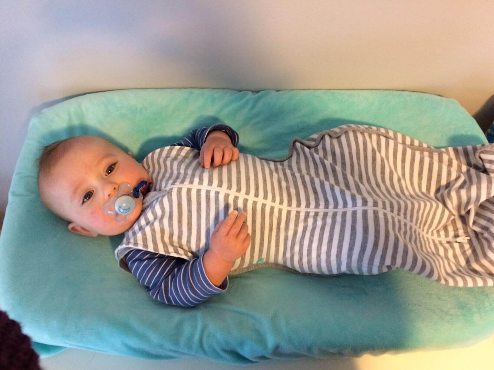 A baby lies on a changing pad covered with a blue blanket. The baby is wearing a grey and white striped Zipadee Zip sleep sack over a blue and white striped onesie, and has a pacifier in their mouth. The baby is looking up, with one hand resting on the chest and the other by the side.