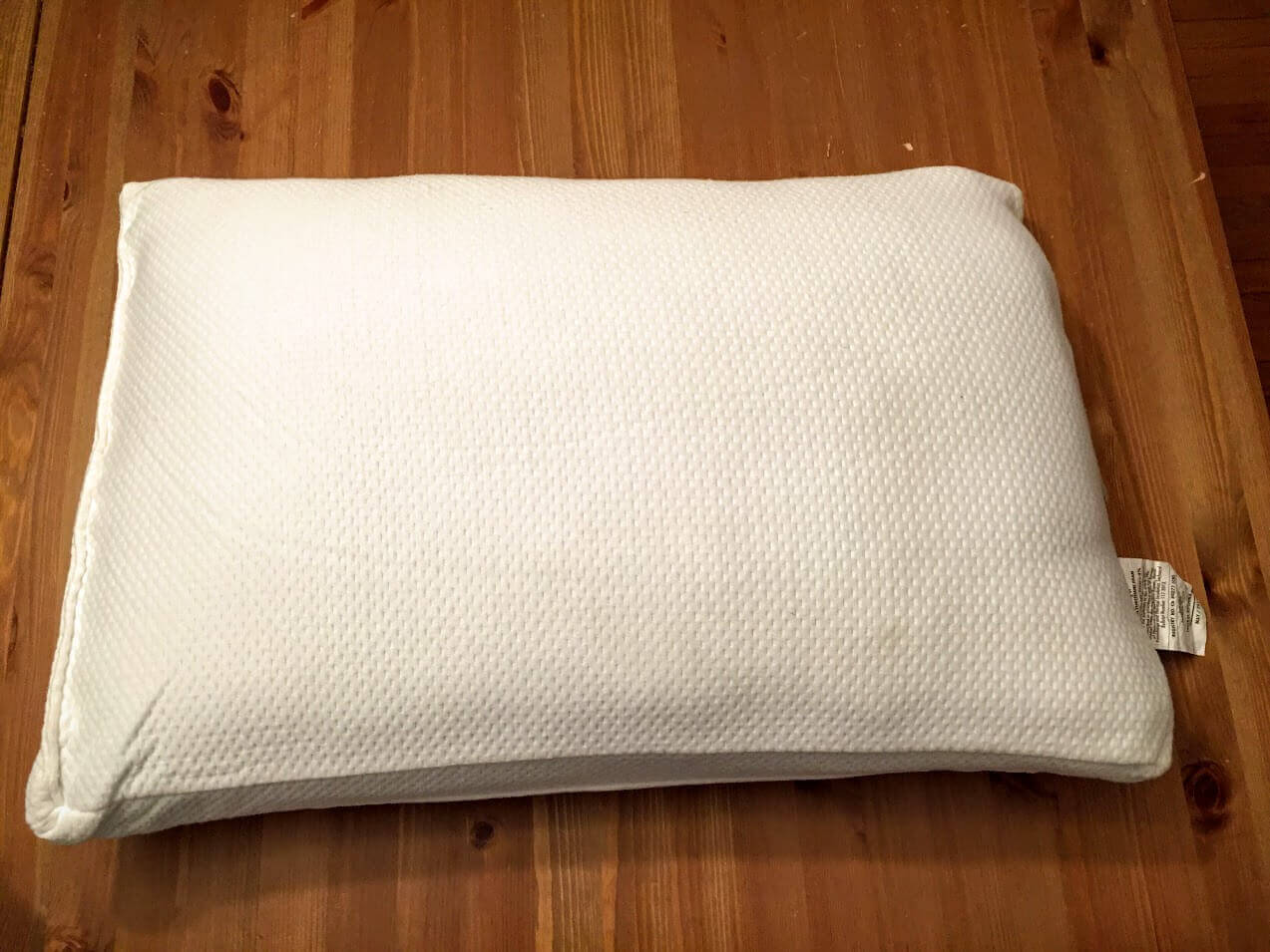 A white Joy Mangano Pillow with a textured cover sits on a wooden surface. The pillow has a small fabric tag on one side.