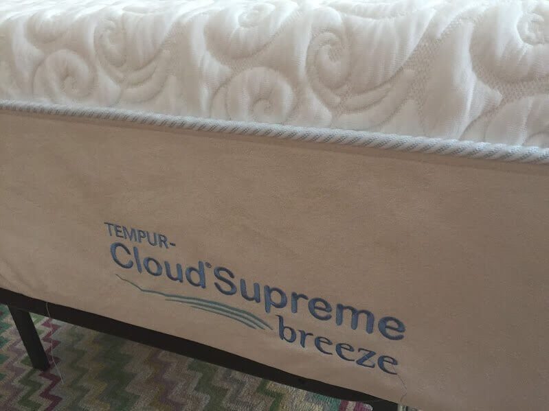 Close-up of the lower side of a mattress showing the brand name "TEMPUR-Cloud Supreme Breeze" stitched in blue on a beige fabric. The Tempurpedic Cloud Supreme Breeze mattress sits on a foundation with a zigzag-patterned carpet partially visible underneath.