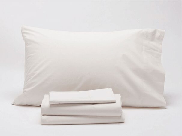 A neatly arranged set of Coyuchi white bed linens, including a large pillow positioned upright behind a folded fitted sheet and a folded flat sheet, all placed on a plain white surface.