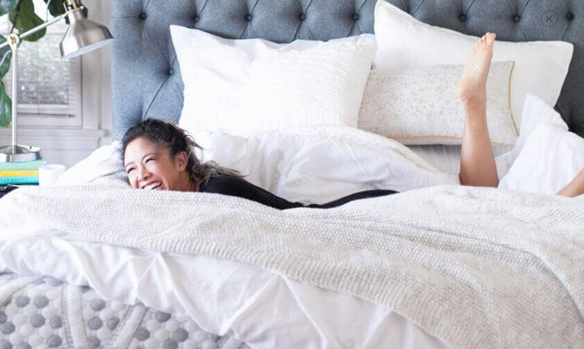 A person lies on a Crave Mattress, smiling and resting on plush white bedding with their legs playfully kicked up. A modern lamp, books, and greenery are visible on the bedside table, adding a cozy and inviting atmosphere to the room.