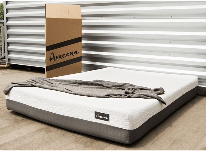 A neatly made Ameena mattress with a white top and gray sides lies on a wooden floor outdoors, accompanied by a gray blanket draped casually over it. A large cardboard box bearing the same brand stands against a corrugated metal wall in the background.