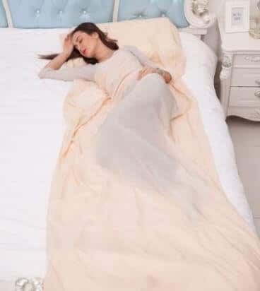 A woman with long dark hair is lying on her back on a bed, covered with a light peach blanket. She has one arm resting on her forehead and the other by her side. The bed has a tufted, light blue headboard and the room appears to be furnished in a white, elegant style.