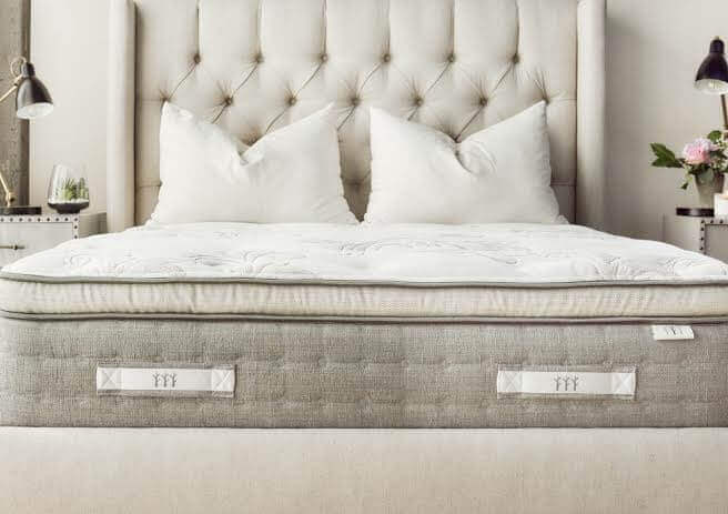 A luxurious, king-sized bed with a tufted headboard and a plush mattress decorated with white pillows. Beside the bed are nightstands with elegant lamps and a small flower arrangement, creating a cozy and inviting bedroom ambiance.