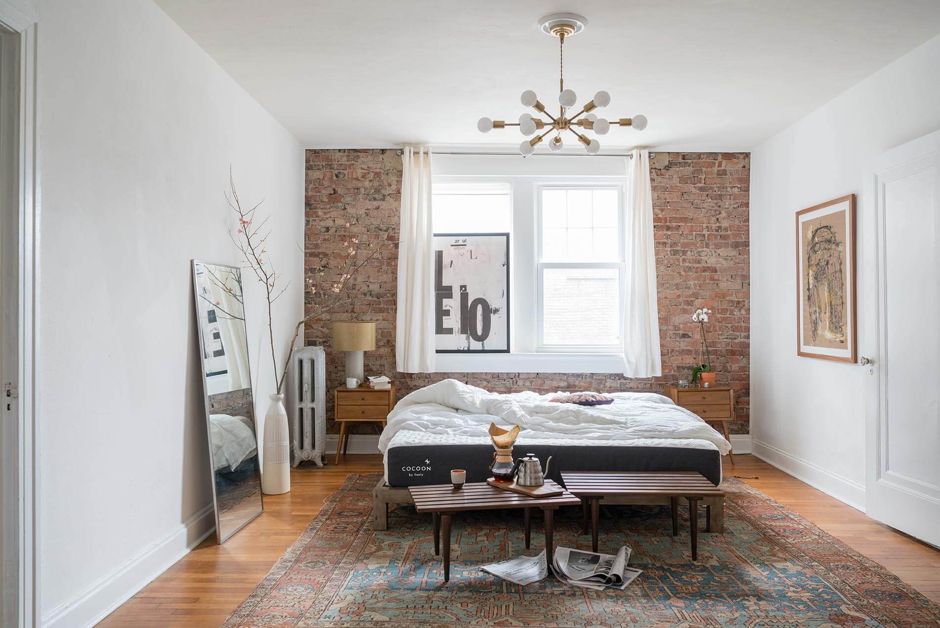 A cozy bedroom with a brick accent wall, a large window with white curtains, and a bed featuring Cocoon Mattress with white bedding. Wooden side tables complement the space, while a decorative chandelier hangs from the ceiling. The room includes a large floor mirror, potted branch decor, and a colorful rug.