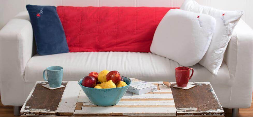 A cozy living room features a white sofa with a red throw and white pillows, including an "I Love My Pillow" accent. A rustic coffee table holds a blue bowl filled with apples and lemons, alongside two mugs, one turquoise and one red, on coasters. A book lies next to the bowl.