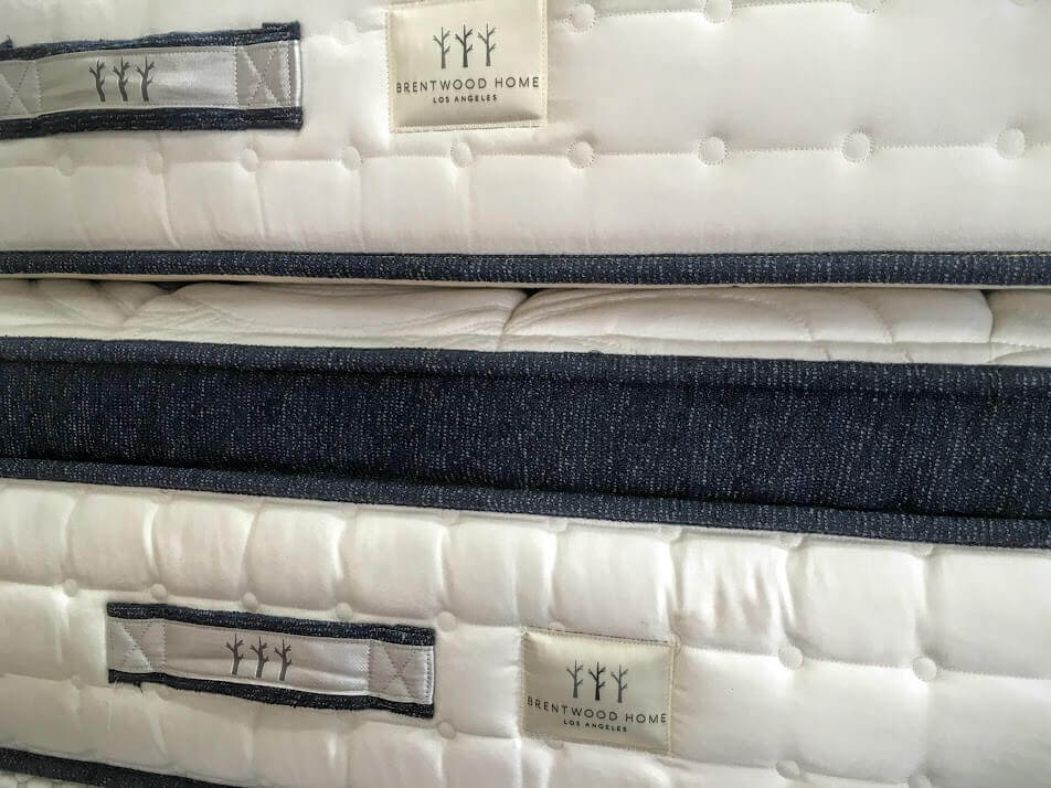 Close-up of the sides of two stacked Brentwood Home mattresses with white quilted fabric and dark blue trim. The fabric labels display the brand name and a simple tree logo. Woven handles are visible on the sides of each mattress.