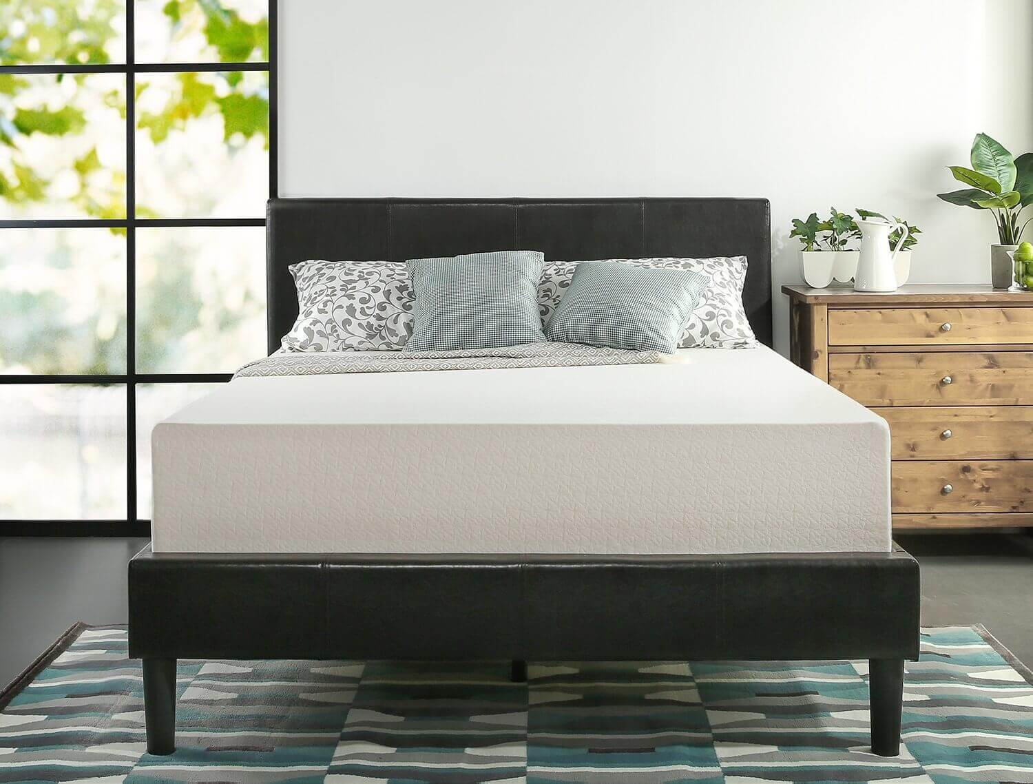 A modern bedroom with a minimalist design. A bed with a white Zinus mattress and gray pillows, including two printed ones, is centered against a window with a leafy view. A wooden dresser with potted plants is on the right. The floor has a geometric-patterned rug.