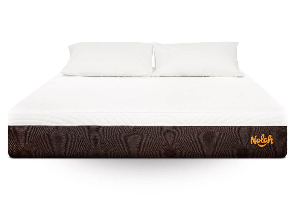 A Nolah Mattress with a white mattress and two white pillows sits atop a brown base featuring a textured pattern. The brand name "Nolah" is visible in orange on the bottom right corner. The bedding appears clean and neatly arranged.
