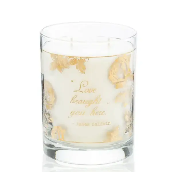 The 22K Gold James Baldwin "Love" Glass Candle - Love Fragrance features a white candle housed in a transparent glass container adorned with elegant golden designs and text. The front of the glass reads, "Love brought you here." - James Baldwin. This candle boasts two wicks and a pristine, smooth surface.