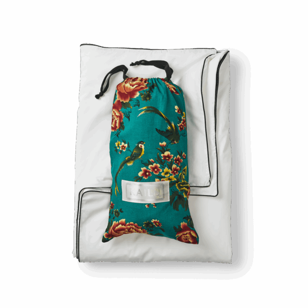 A white pillow with black piping along the edges is laid flat. On top of it, there is a drawstring bag made of teal fabric adorned with a vibrant floral pattern featuring red and yellow flowers and green leaves. The bag, part of the Open Road Portable Silk Throw Set, has a clear label with text on it.