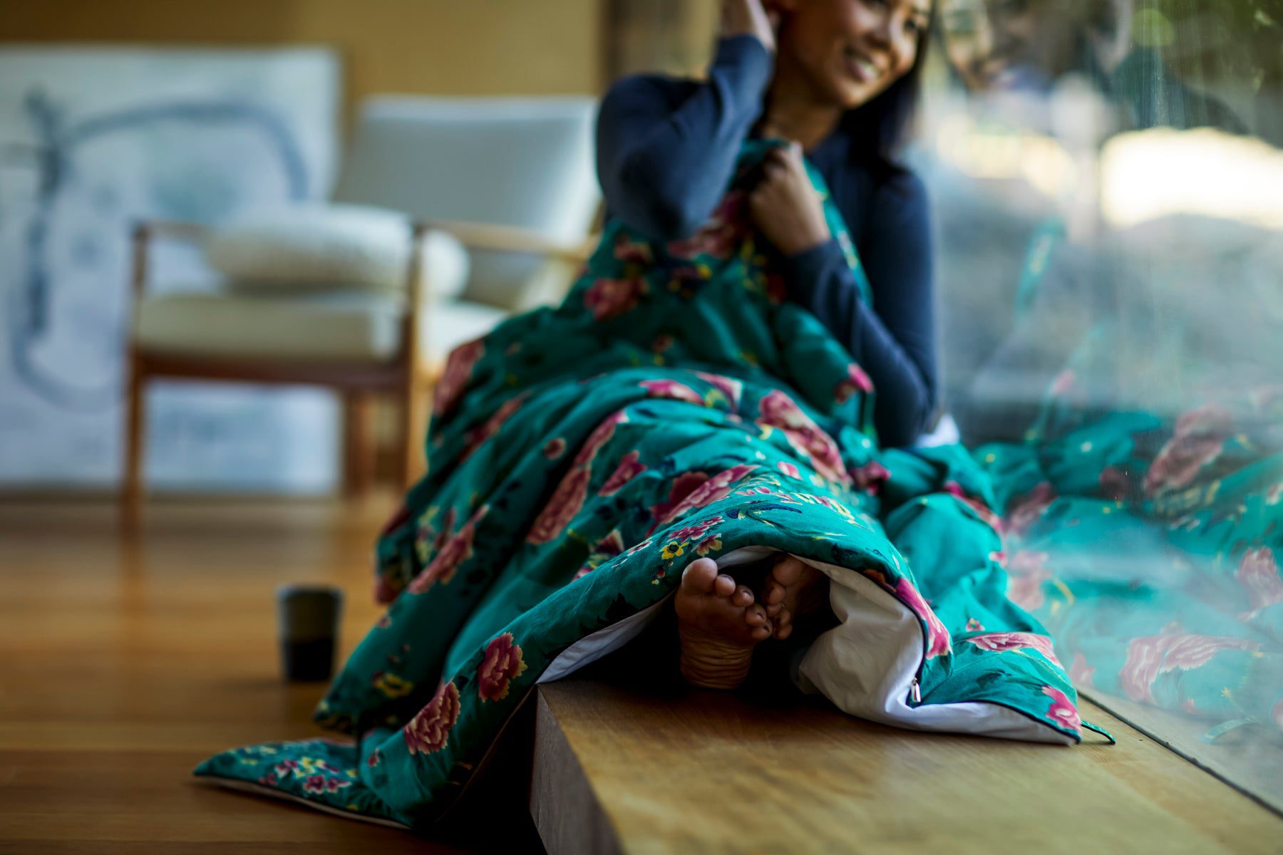 A person sits by a large window, wrapped in an Open Road Portable Silk Throw Set. They have one hand behind their head and appear to be smiling. The person's barefoot toes are peeking from under the silk throw. A mug sits on the wooden floor beside them.