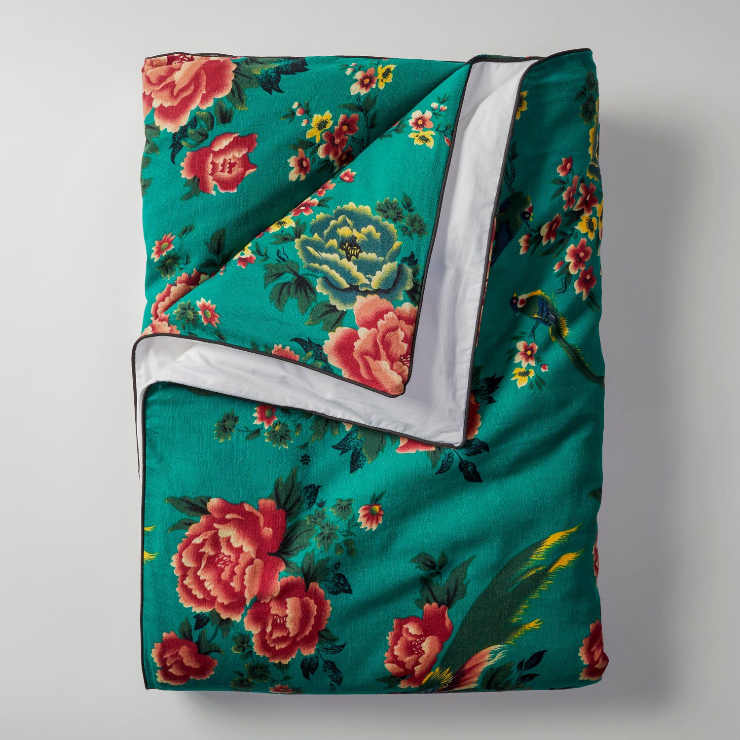 The Open Road Portable Silk Throw Set showcases a vibrant teal-colored quilt adorned with a floral pattern featuring large red, pink, and yellow flowers with green leaves. The quilt is partially folded to reveal its white underside and is displayed against a plain white background.
