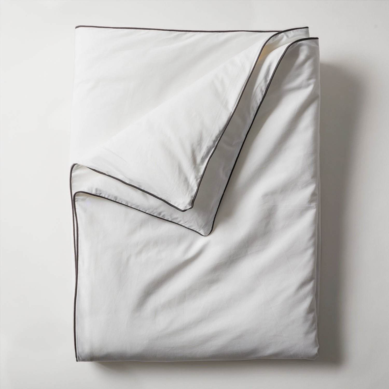 An elegantly folded Open Road Portable Silk Throw Set in a pristine white shade, featuring refined dark piping along its edges, rests on a light-colored surface. The top edge is gently turned down, showcasing the interior side. The overall look is clean and minimalistic.