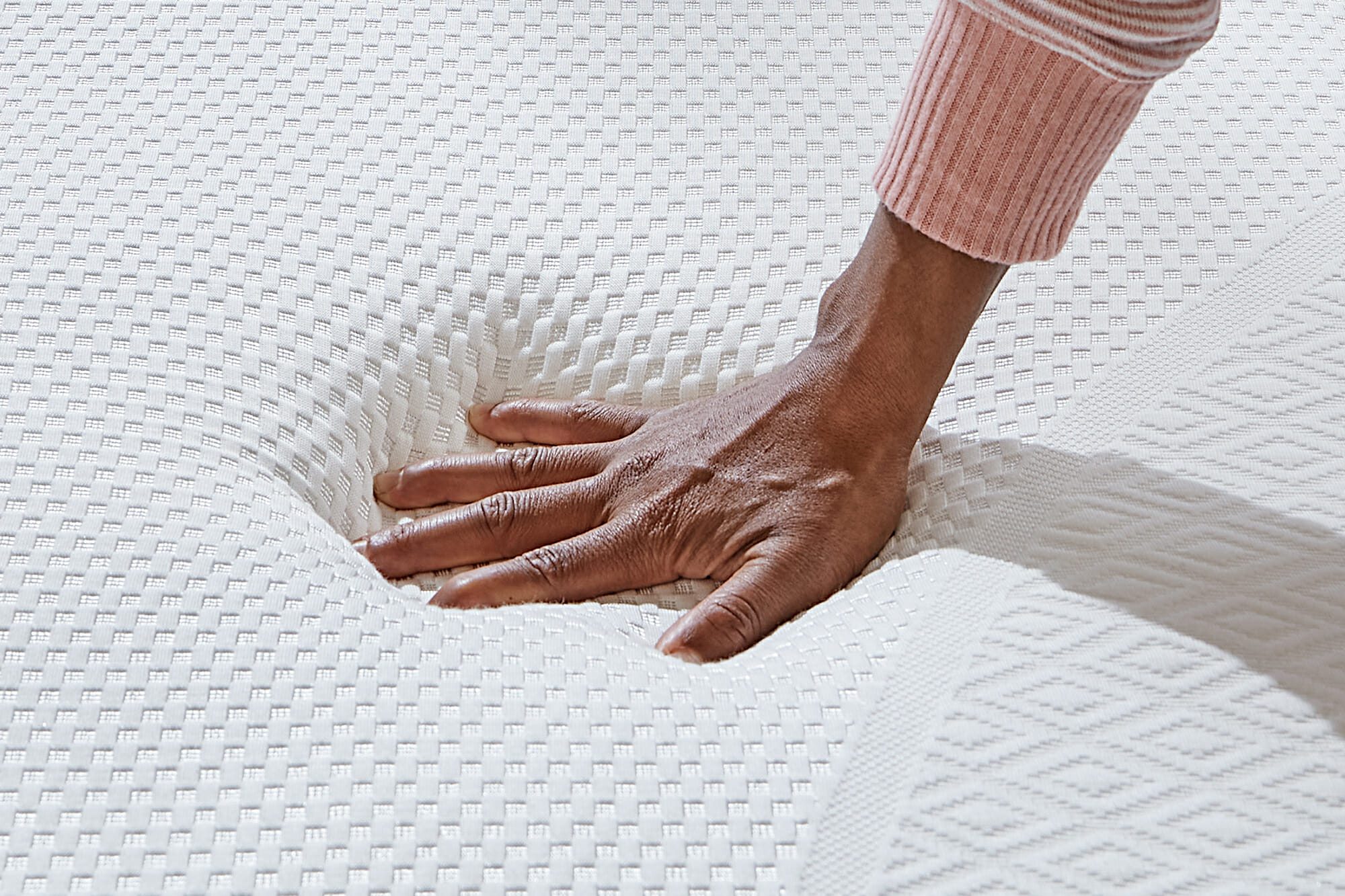 A hand pressing down on the Mlily Dream white textured mattress, demonstrating its softness and comfort. The person is wearing a long-sleeved pink top with white stripes.