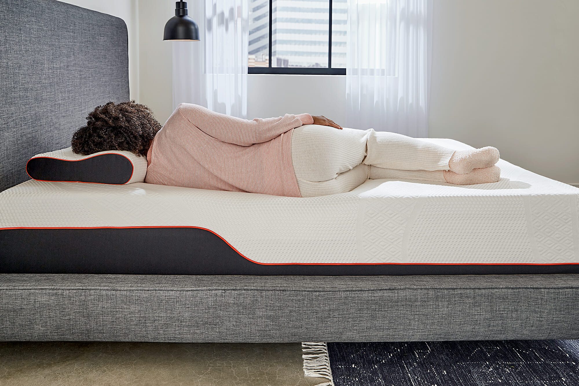 A person is lying on their side on a {{Product Name: Mlily Dream}} mattress, covered by a pink top and white pants. The room features a hanging black light fixture, a large window with sheer white curtains, and a gray bed frame with a black and white rug on the floor.