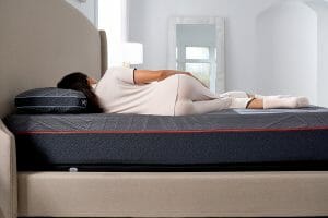 A person is lying on their side on the Mlily PowerCool Medium Mattress, which features a modern, dark-colored design. They are wearing light-colored pajamas and socks. The bed is complemented by a beige upholstered headboard, while a large mirror and a lamp can be seen in the bright room's background.