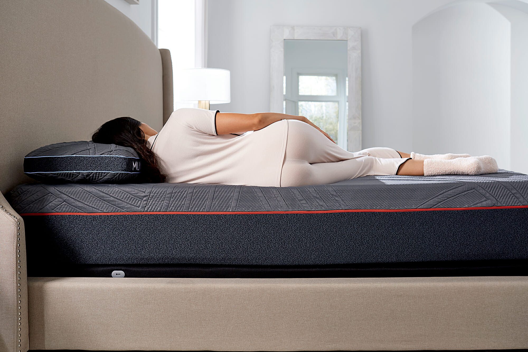 A person is lying on their side on the Mlily PowerCool Medium Mattress, which features a modern, dark-colored design. They are wearing light-colored pajamas and socks. The bed is complemented by a beige upholstered headboard, while a large mirror and a lamp can be seen in the bright room's background.